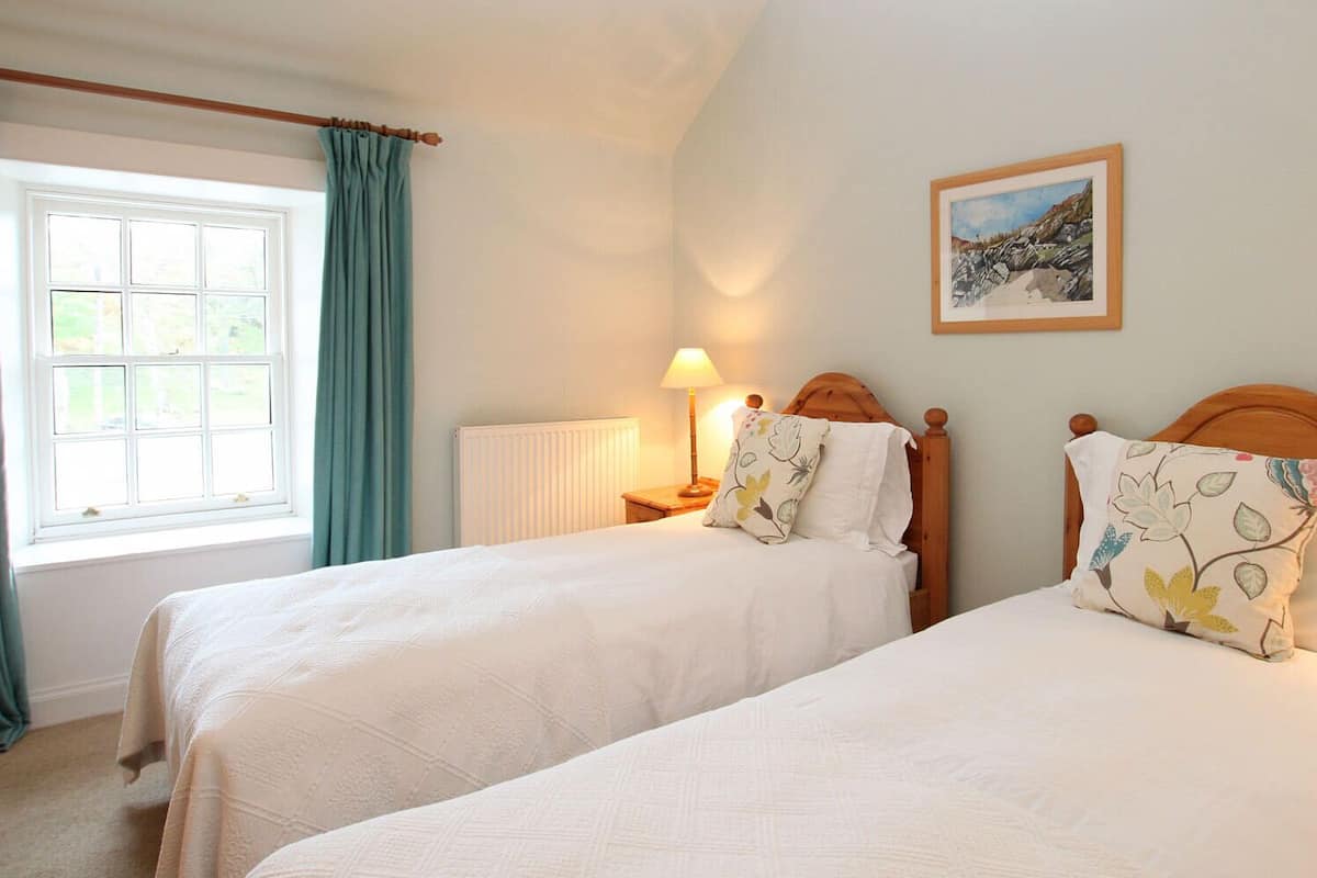 A bedroom with two single beds, a window with curtains, a bedside table with a lamp, and a framed picture on the wall.