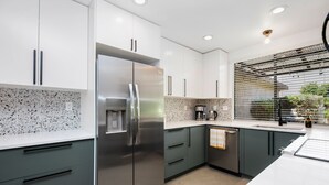 A modern kitchen with white cabinets, a stainless steel refrigerator, and a window with blinds. - Winter with us in beautiful sunny Palm Springs! (Palm Springs)