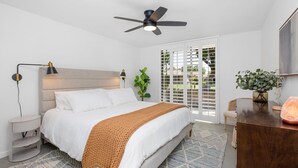 A modern bedroom with a bed, bedside tables, a ceiling fan, a window with blinds, a plant, and a desk with a lamp. - Winter with us in beautiful sunny Palm Springs! (Palm Springs)