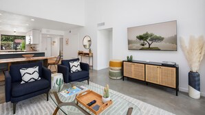 A modern living room with a glass coffee table, two sofas, a dining area with a kitchen in the background, and a large painting of a tree on the wall. - Winter with us in beautiful sunny Palm Springs! (Palm Springs)