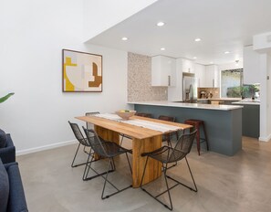 A modern kitchen with a central island, a dining area with a wooden table and wire mesh chairs, and a kitchen counter with a coffee machine.