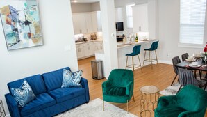Ein modernes Wohnzimmer mit einem blauen Sofa, grünen Sesseln und einem Essbereich mit Tisch für vier Personen.