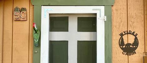 Een houten blokhut met een groene deur en een raam. Op de muur staat het cijfer '50' en een logo.