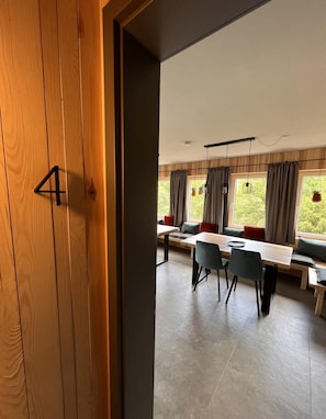 Ein moderner Essbereich mit einer Holztür, auf der '4' steht, einem langen Tisch mit Stühlen und großen Fenstern mit Vorhängen.