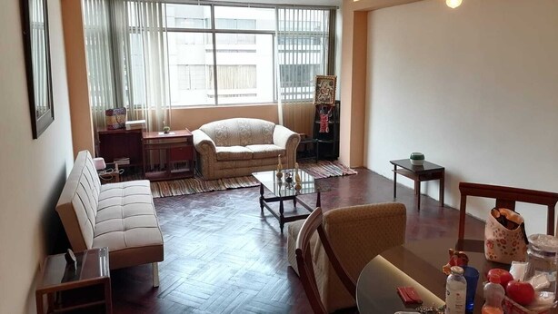 A living room with a sofa, armchairs, a coffee table, and a dining table. There is a large window with curtains, a chandelier, and a small table with decorative items.