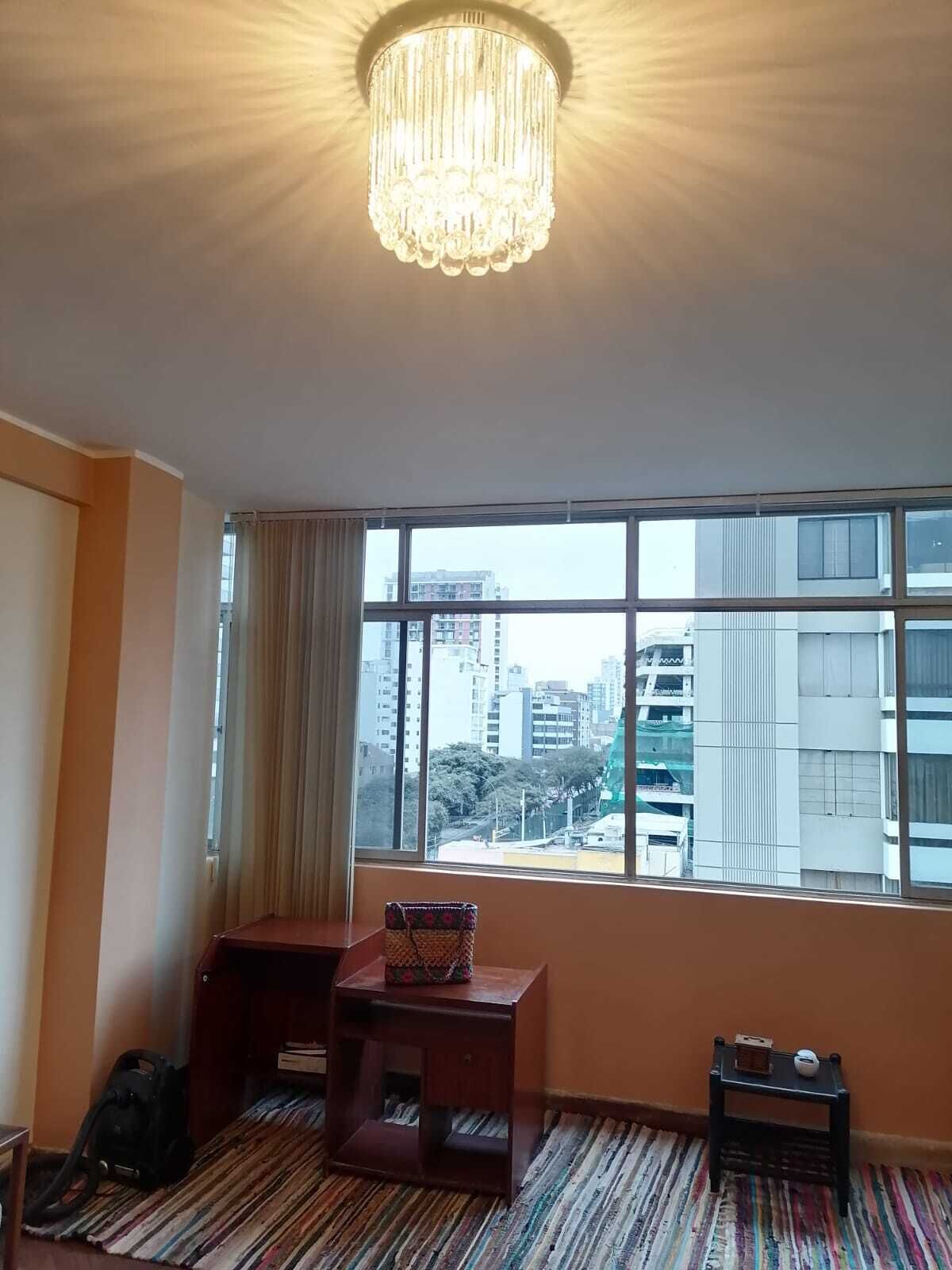 A hotel room with a large window, a chandelier, a small wooden table, a suitcase, and a striped rug.