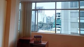 A hotel room with a large window, a chandelier, a small wooden table, a suitcase, and a striped rug.