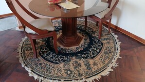 A round glass table with a dark wooden base, surrounded by two wooden chairs with cushioned seats. A decorative rug with a floral pattern is under the table.