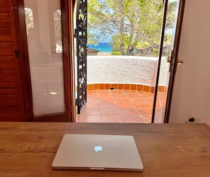 A laptop on a wooden table, a door with a metal gate, and a view of a tiled patio with a stone wall and trees.