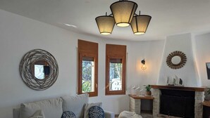 A cozy living room with a sofa, ottoman, and a fireplace. There are two windows with wooden shutters, a wall-mounted mirror, and a hanging light fixture. - 1 minute to the beach! Family-sized Garden and BBQ (Miami Platja)