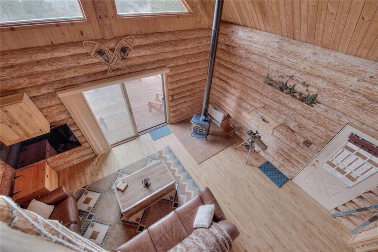 A wooden cabin interior with a loft bed, a fireplace, and a large window.