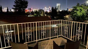 Un balcon doté d'une balustrade en métal, de chaises en osier et offrant une vue sur les toits d'une ville illuminée la nuit.