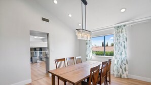 Un coin repas avec une table et des chaises en bois, un lustre suspendu et une vue sur un jardin, visible à travers une fenêtre ornée de rideaux à motifs.