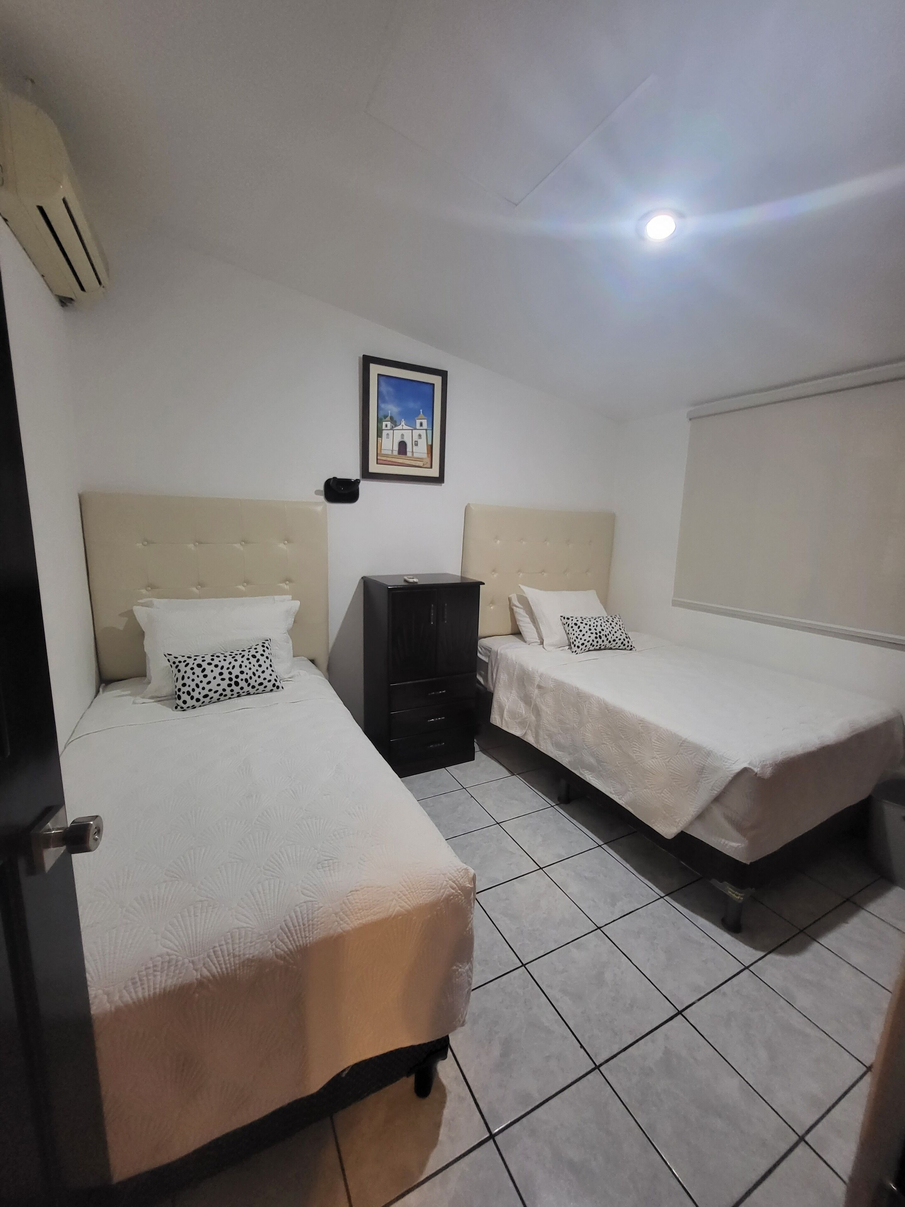 A hotel room with two beds, a small black cabinet, a framed picture on the wall, and a ceiling light.