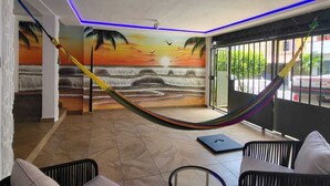 Ein Zimmer mit einer Wandmalerei, die einen Sonnenuntergang über Wasser zeigt, einer Hängematte und zwei schwarzen Korbstühle mit Kissen.