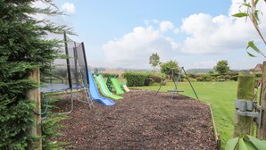 A playground with slides and swings, surrounded by a grassy area and trees. - 10 Old Orchard (Much Wenlock)