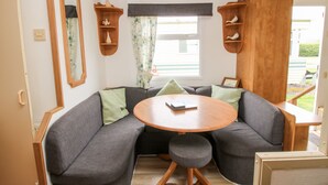 A cozy living area with a curved grey sofa, a round wooden table, and a small stool. There are wooden shelves with decorative items and a window with curtains. - 6 Old Orchard (Much Wenlock)