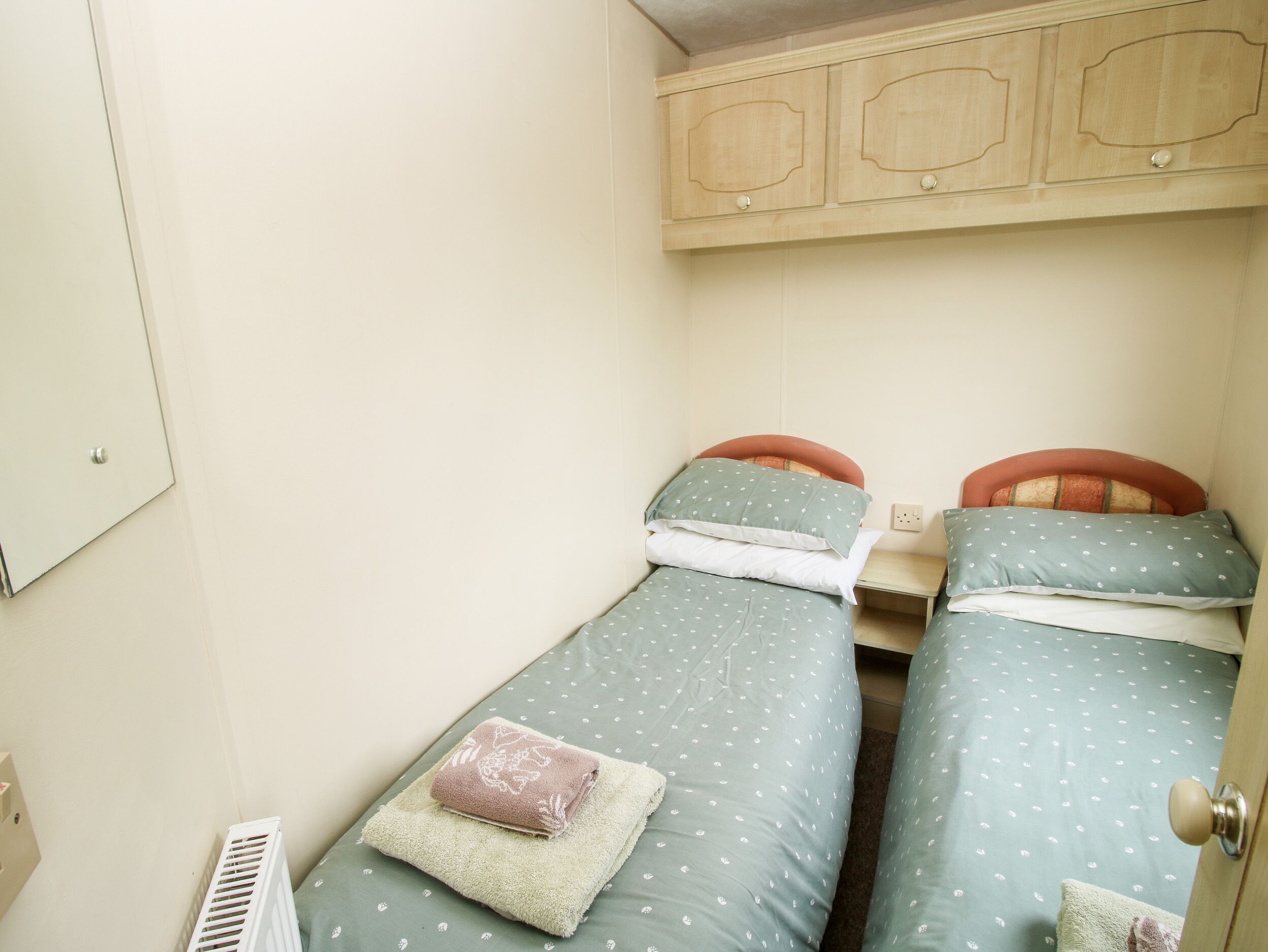 A small, single-room accommodation with two beds, each with a headboard and pillows. There is a wooden cabinet above the beds and a radiator on the floor.