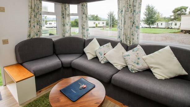 A corner sofa with floral cushions, a small wooden side table, and a view of a caravan park. - 6 Old Orchard (Much Wenlock)