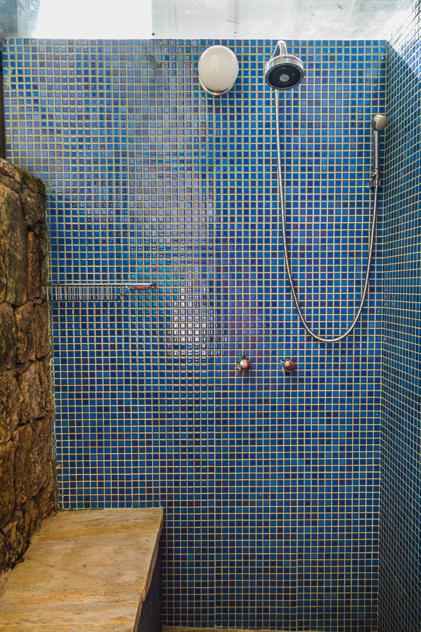 En dusch med blå mosaikplattor, en vägg av natursten och ett duschmunstycke i metall.