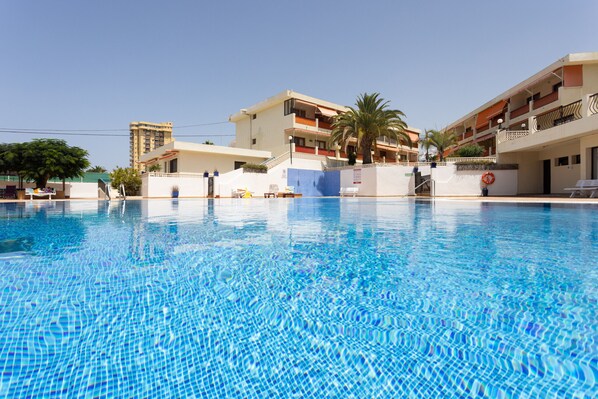 Una piscina con agua cristalina azul, rodeada de edificios con balcones y una palmera.