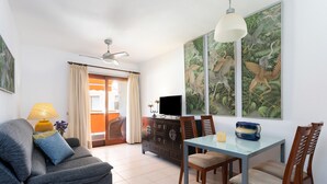 Een woonkamer met een bank, een eettafel met stoelen, een televisie, een kast en een plafondventilator.