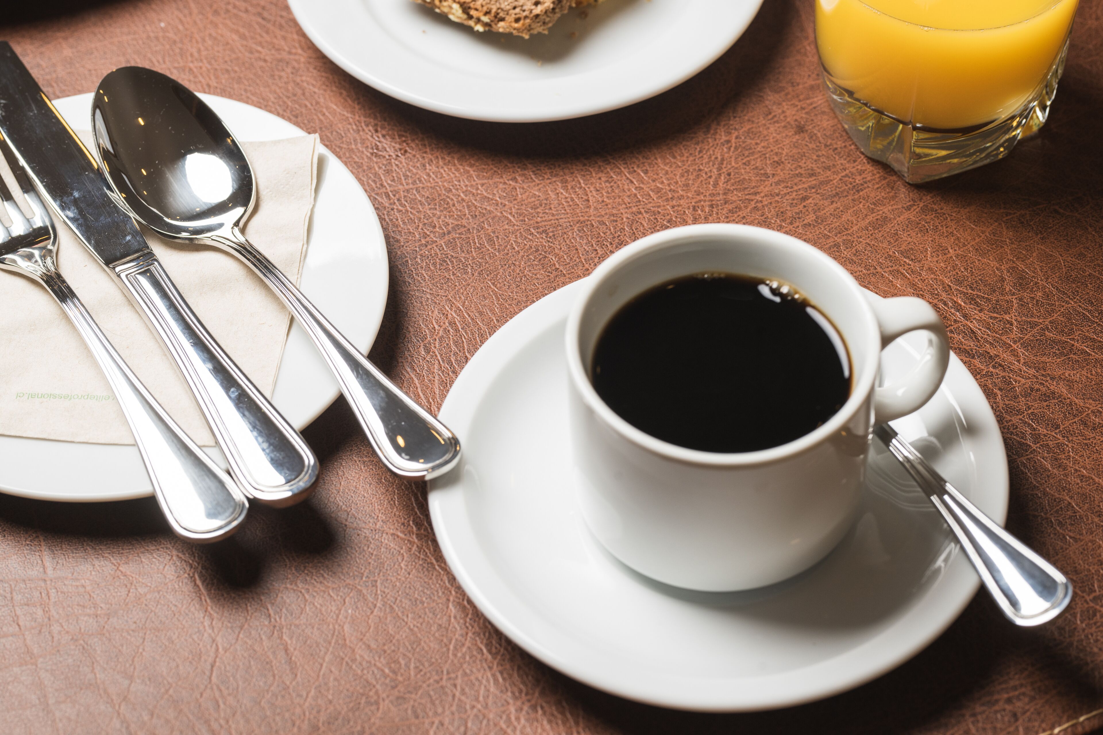 Une table avec une tasse de café noir, un verre de jus d’orange et une assiette avec une tranche de pain.