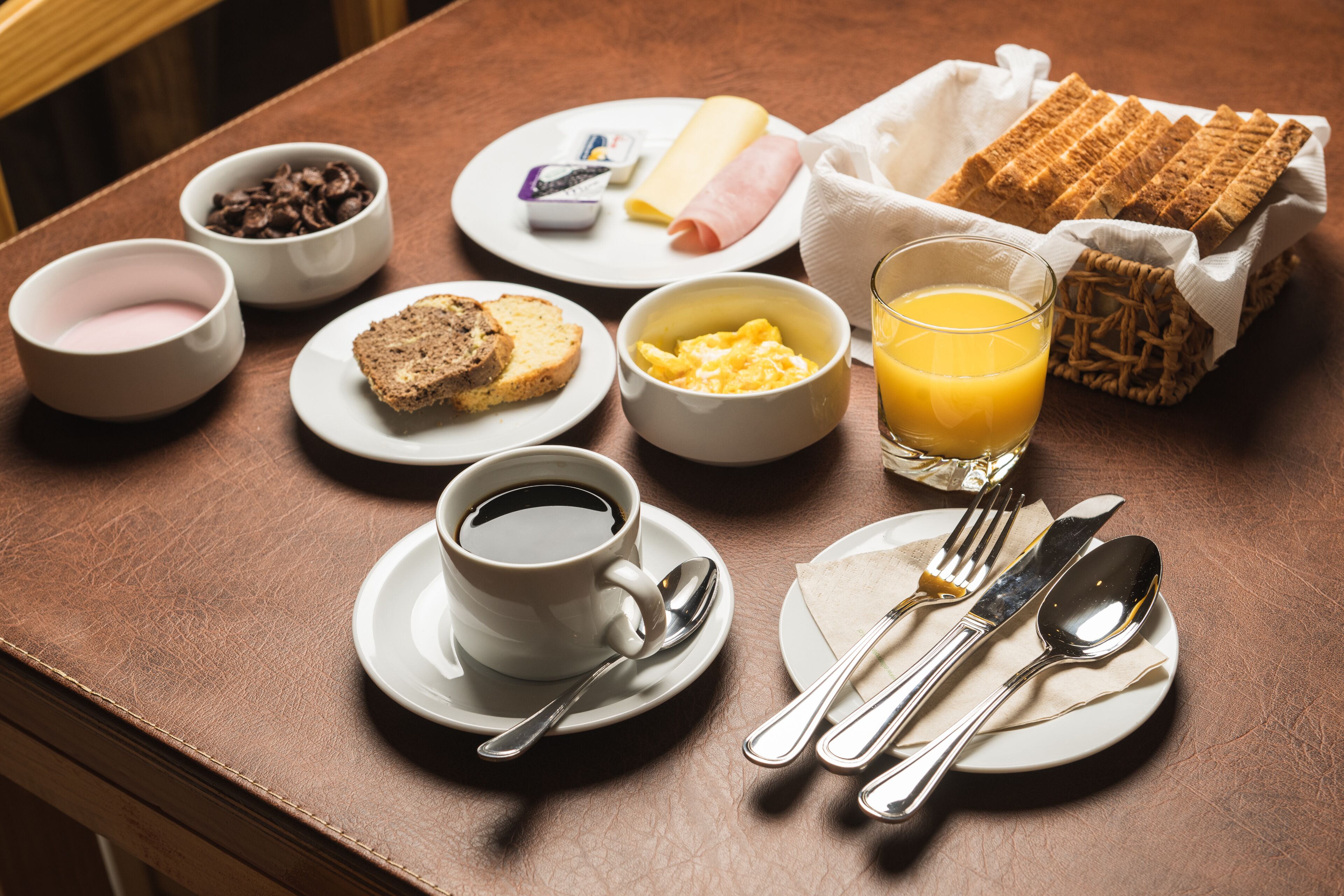Uma mesa de madeira com café, torradas, ovos mexidos, presunto, queijo e um copo de suco de laranja.