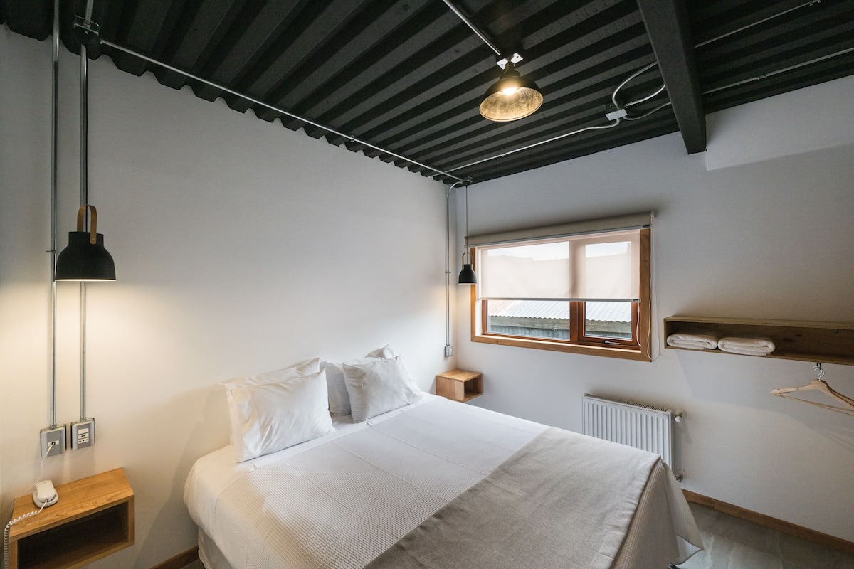 A bedroom with a bed, bedside lamp, wooden nightstand, and a window with a view.