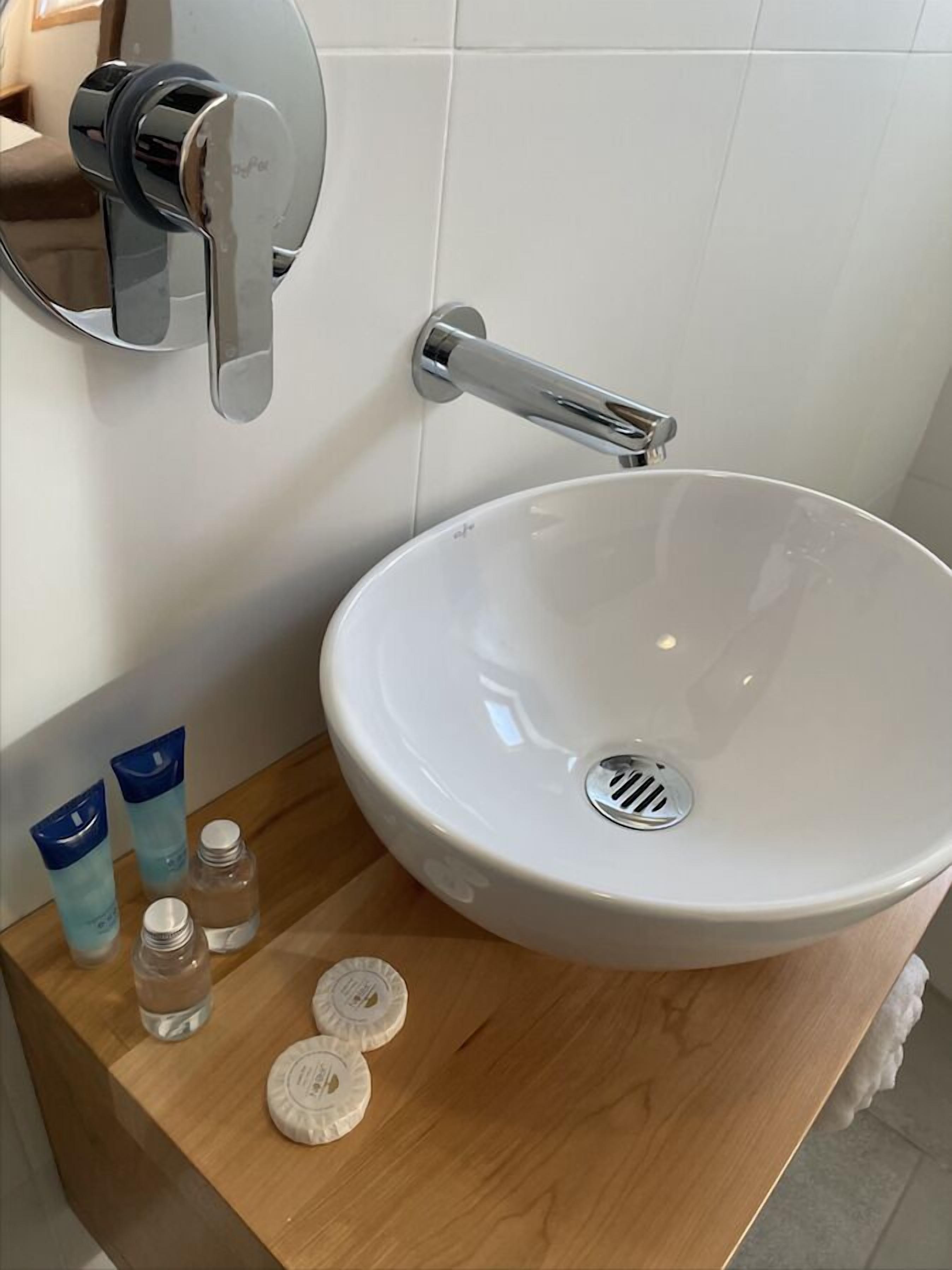 A bathroom with a white sink, a mirror, and toiletries on a wooden shelf.