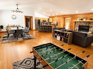 A modern kitchen with wooden cabinets, a dining area with a table and chairs, a living room with a sofa and a fireplace, and a foosball table.