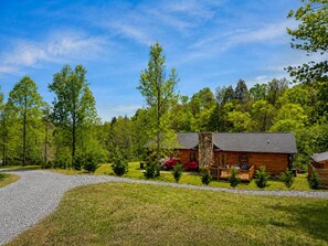 A cabin with a stone chimney, surrounded by a gravel driveway and lush greenery.