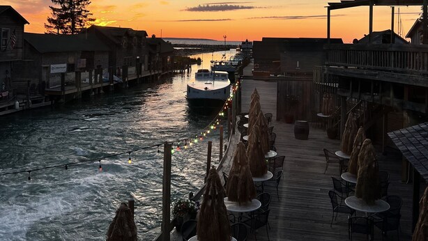 Restaurante à beira-rio com mesas ao ar livre, oferecendo vista para o rio, um barco atracado nas proximidades e um pôr do sol ao fundo.