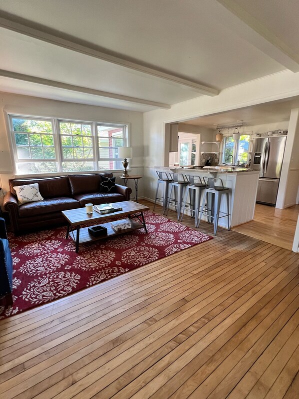 A modern living room with a wooden floor, a red patterned rug, a brown leather sofa, and a dining area with bar stools.