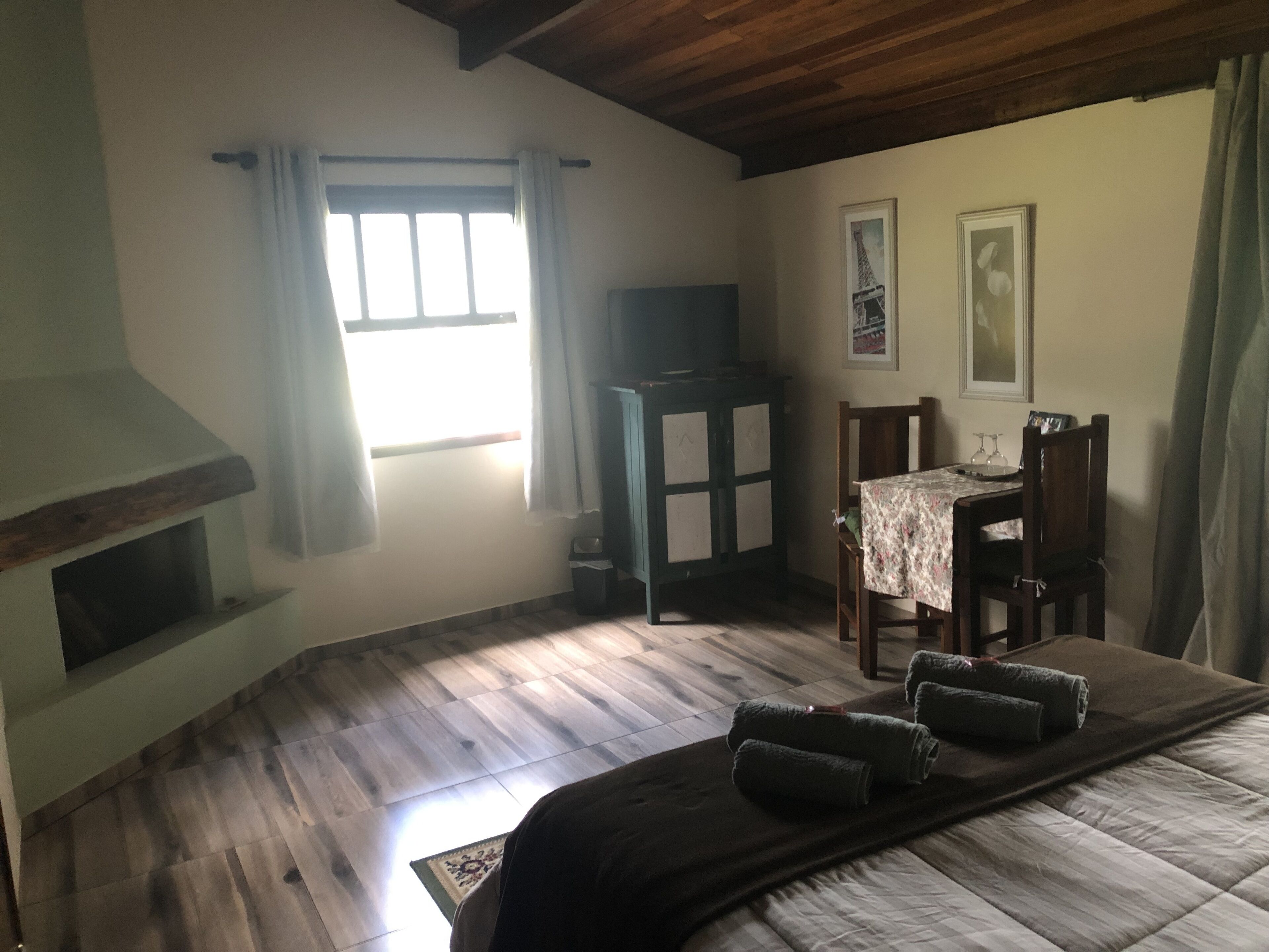 Una habitación acogedora con una cama, una mesita con una silla, un televisor y un ventanal con cortinas.
