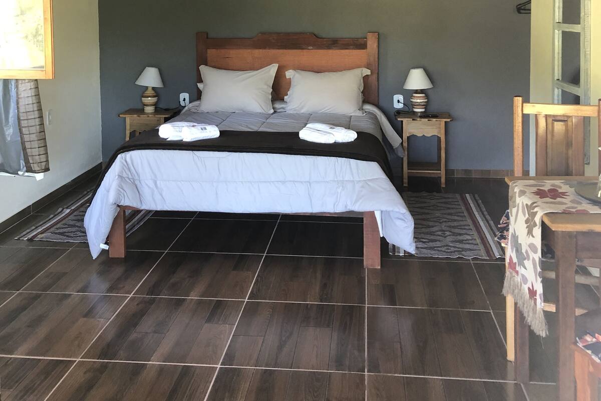Un dormitorio con techo de madera, una cama con ropa de cama blanca, dos mesitas de noche con lámparas y una pequeña mesa con mantel floreado.