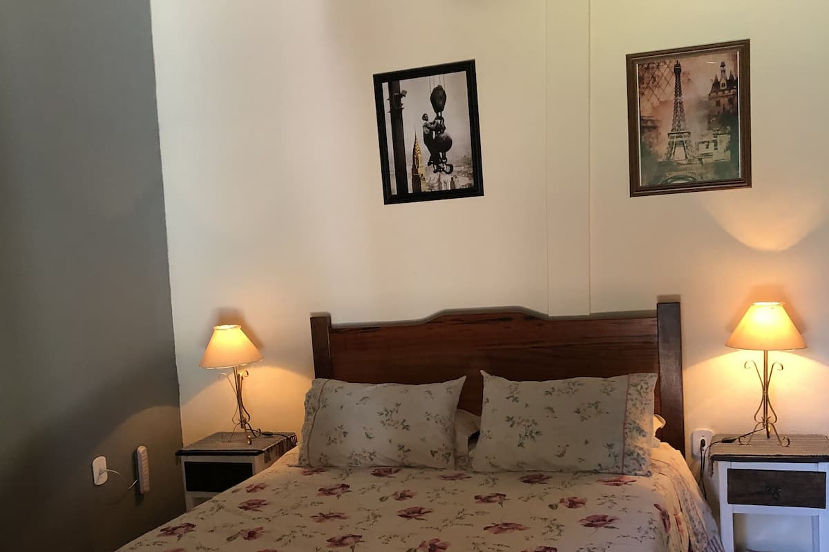 Un dormitorio con cama de madera, ropa de cama floral, dos lámparas de noche, una mesita de noche, un cuadro enmarcado con una vista urbana y una lámpara colgante.