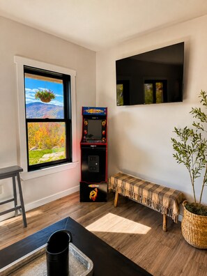 Ein Zimmer mit Fenster, einem an der Wand montierten Fernseher, einer Spielkonsole, einer Bank und einer Topfpflanze.