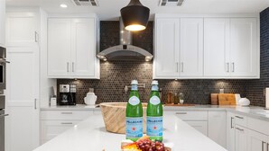 Cocina moderna con armarios blancos, salpicadero negro y una isla central con botellas de agua Pellegrino y fruta.