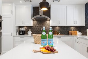A modern kitchen with white cabinets, a black backsplash, and a central island with Pellegrino water bottles and fruit.