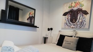 Uma cama bem arrumada com travesseiro com texto, um espelho fixado na parede e uma pintura de uma ovelha.
