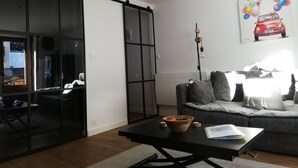 Uma sala de estar moderna com uma porta de vidro deslizante, um sofá cinzento, uma mesa de centro preta com uma tigela e um livro, e um quadro de um carro na parede.