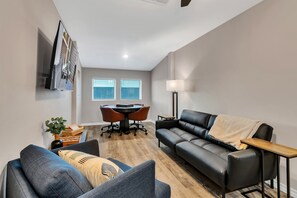 Un salon moderne avec un canapé en cuir noir, un fauteuil gris et une table basse en bois. On y trouve également une table à manger avec des chaises et une lampe dans un coin.