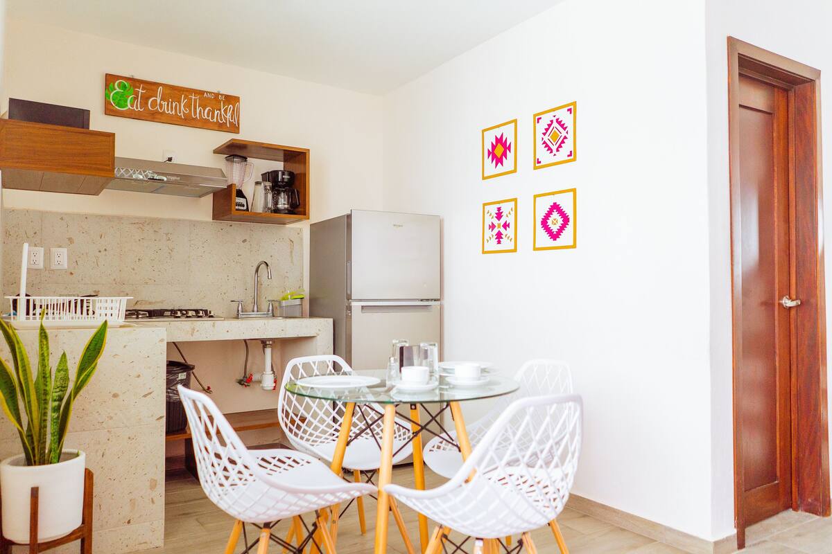 A compact kitchen with a dining area, white chairs, a small refrigerator, and a coffee machine.