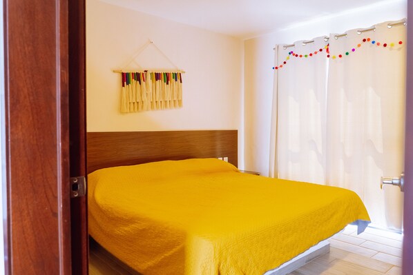 Una habitación con cama, cabecera de madera y cortina con flecos coloridos.