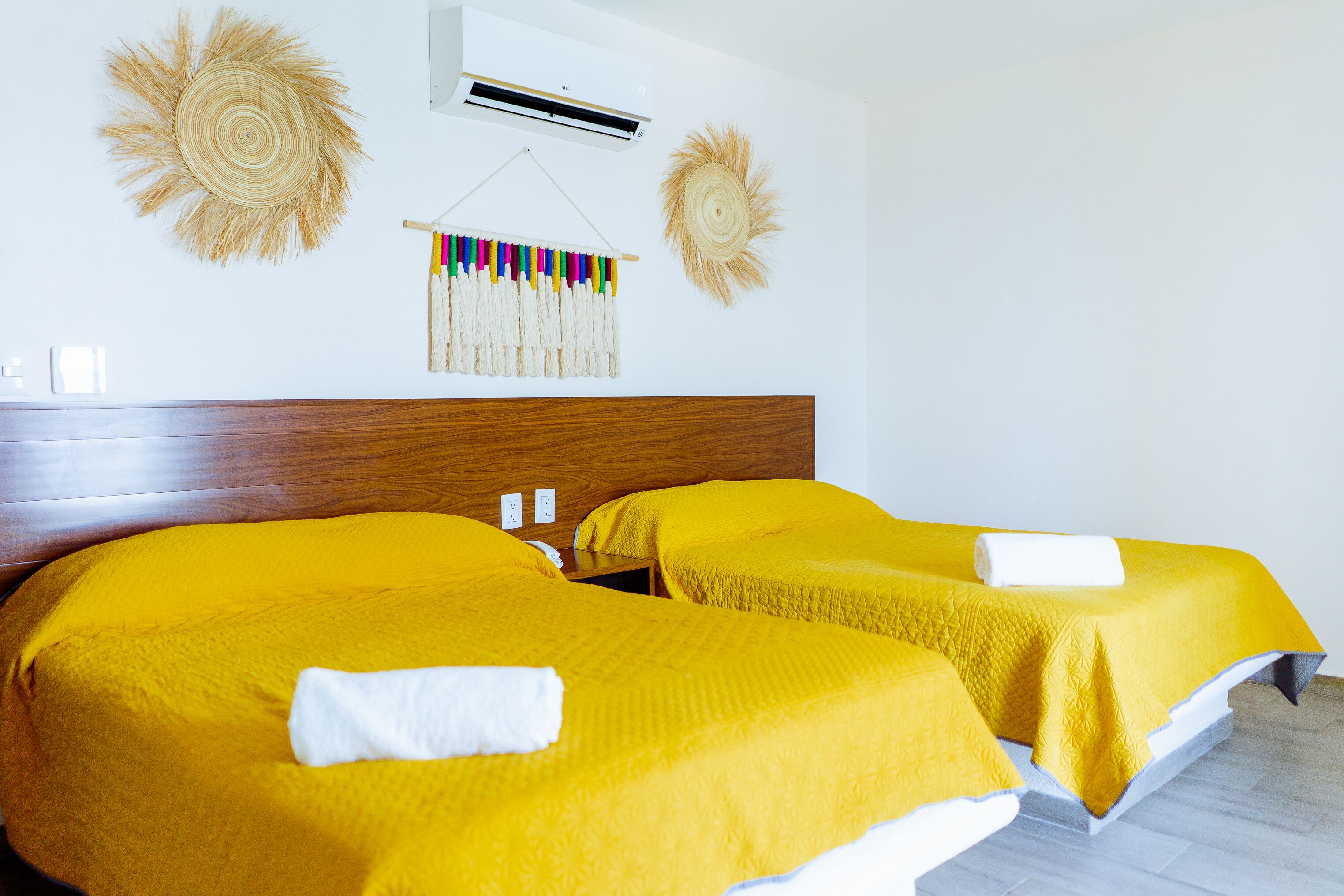 A hotel room with two beds, each with a yellow bedspread and a white towel. There is a wooden headboard, an air conditioning unit, and wall decorations.