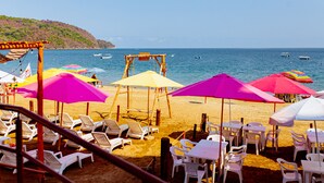 Un área de descanso en la playa con sillas reclinables blancas y mesas, sombrillas de colores, y vistas al mar y a los barcos.