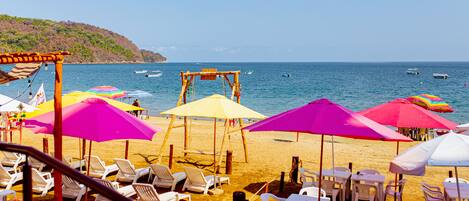 A beachside seating area with white lounge chairs and tables, colorful umbrellas, and a view of the sea and boats.