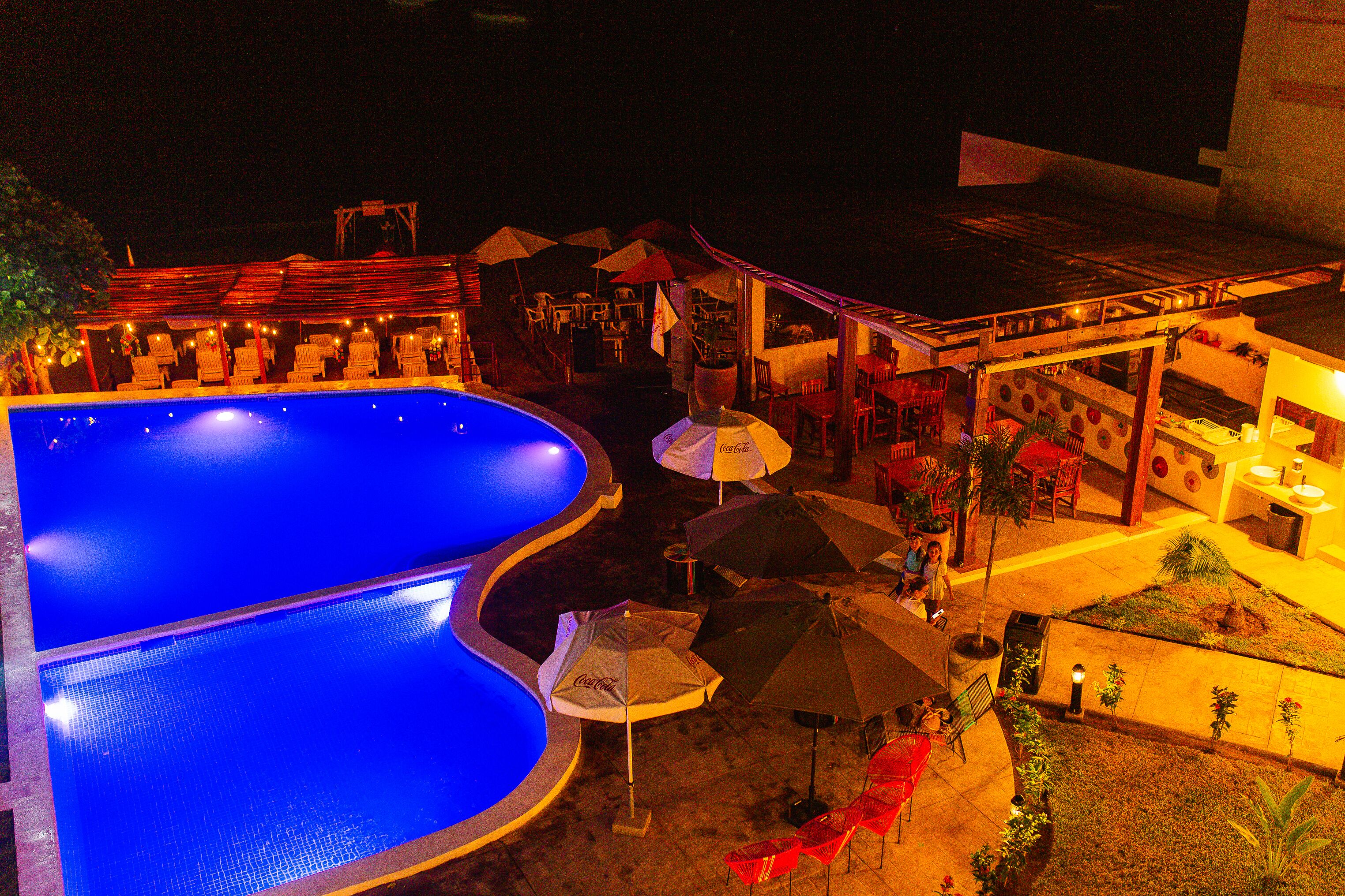 A nighttime view of a pool area with lounge chairs and umbrellas, a bar area with tables and chairs, and a small garden with plants.