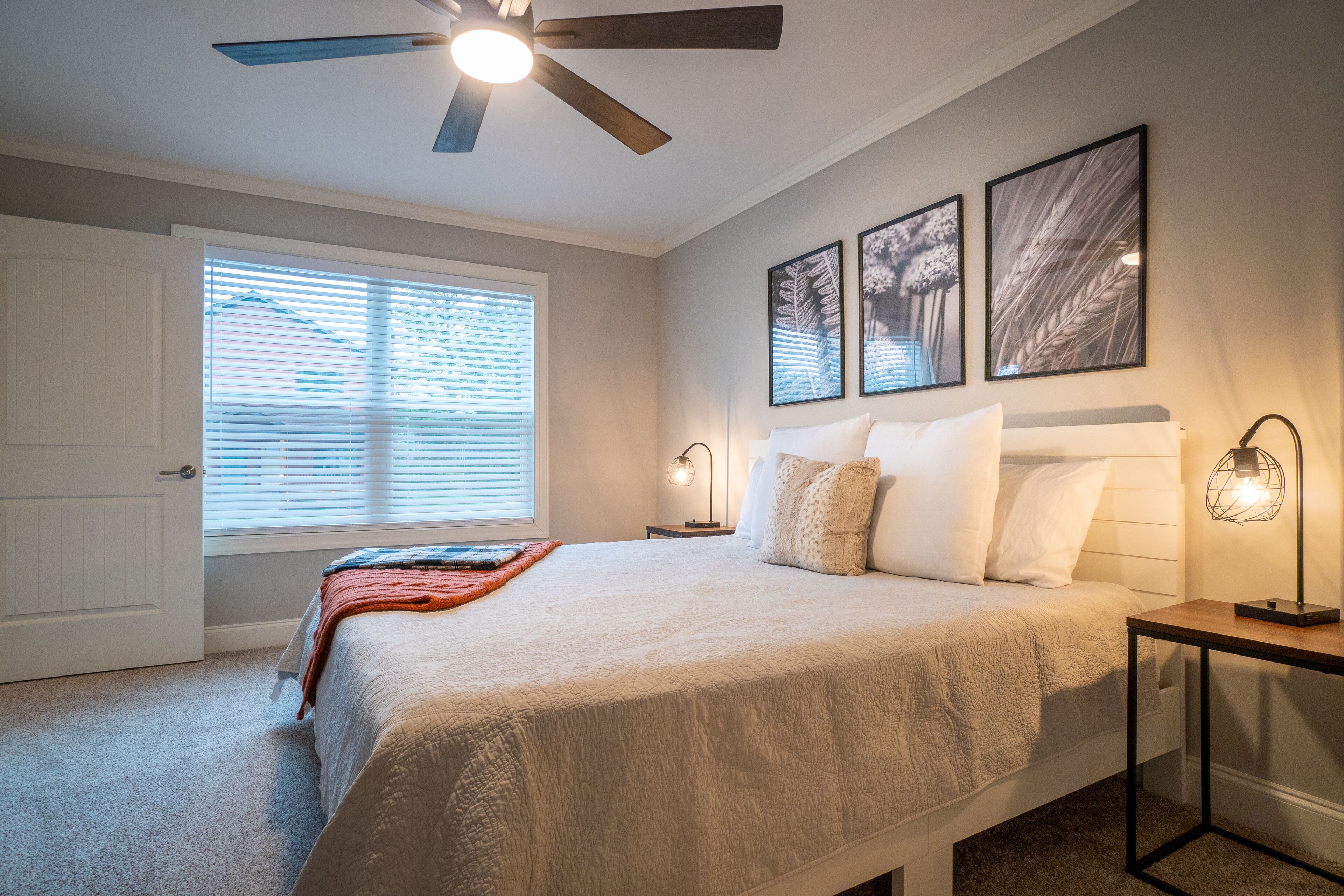 A bedroom with a bed, a ceiling fan, a nightstand, a lamp, and framed pictures on the wall.